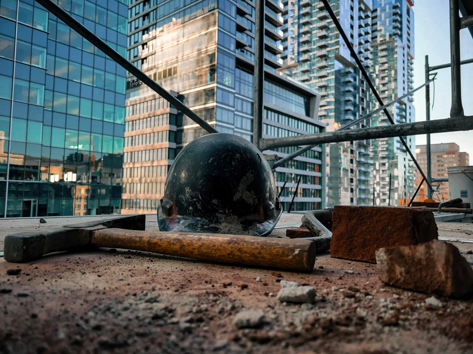 View of a high-rise construction site with a safety helmet, hammer, and bricks, surrounded by modern buildings in the background.