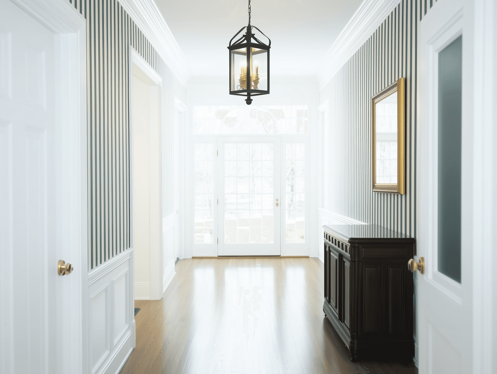 Bright and stylish entryway with striped walls, hanging chandelier, dark wood sideboard and glass doors.