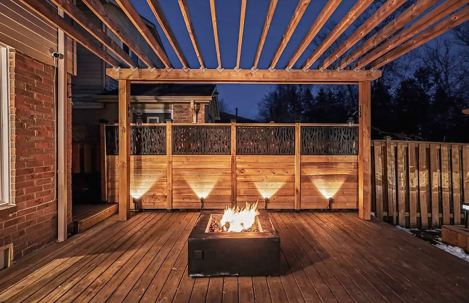 Wooden deck with pergola and illuminated outdoor fireplace, surrounded by a decorative wooden fence, perfect for warm outdoor evenings.