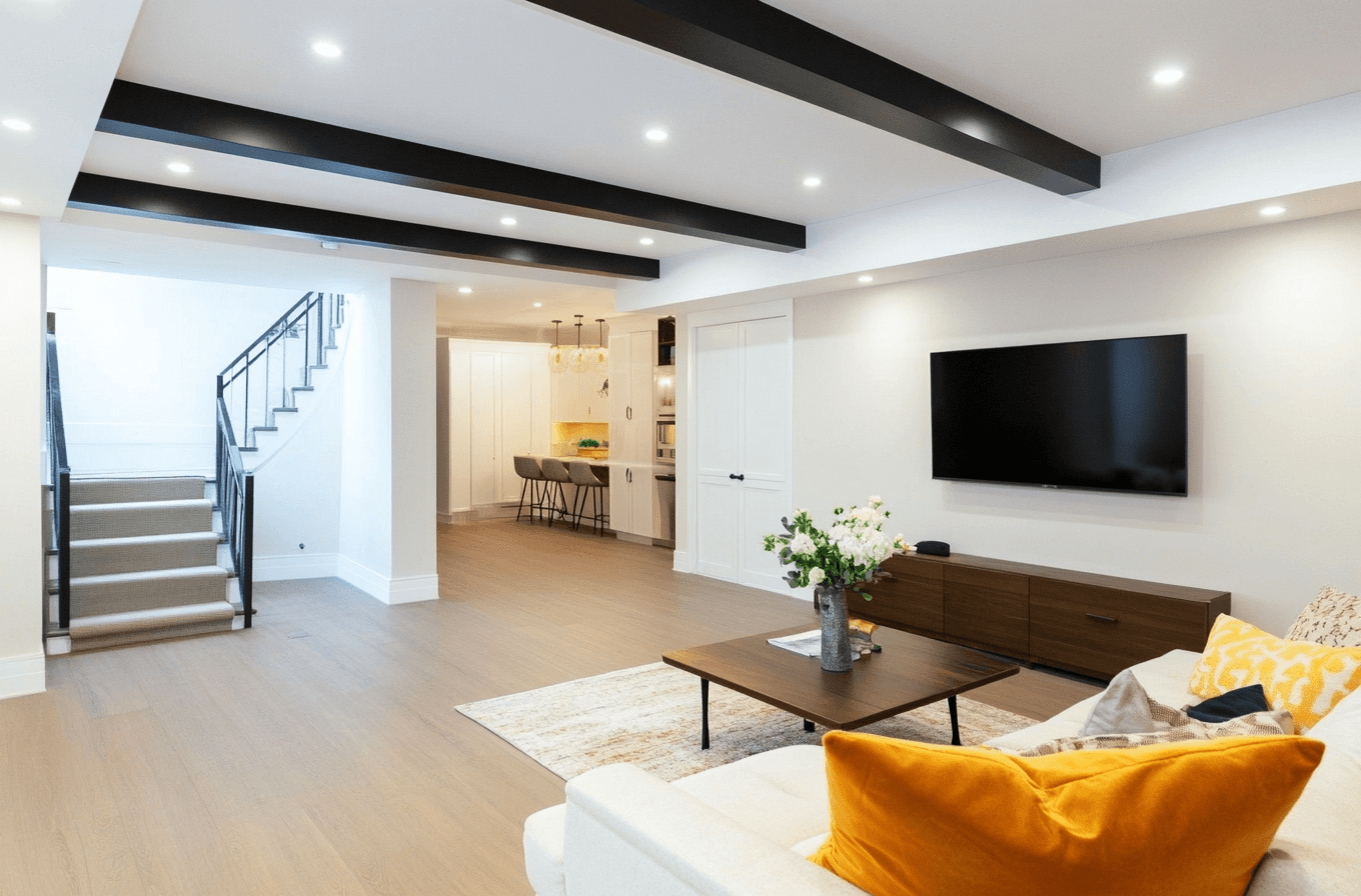 Bright and modern living room with black ceiling beams, white sofa with yellow cushions, wall-mounted TV, elegant staircase, and dining area with pendant lighting.