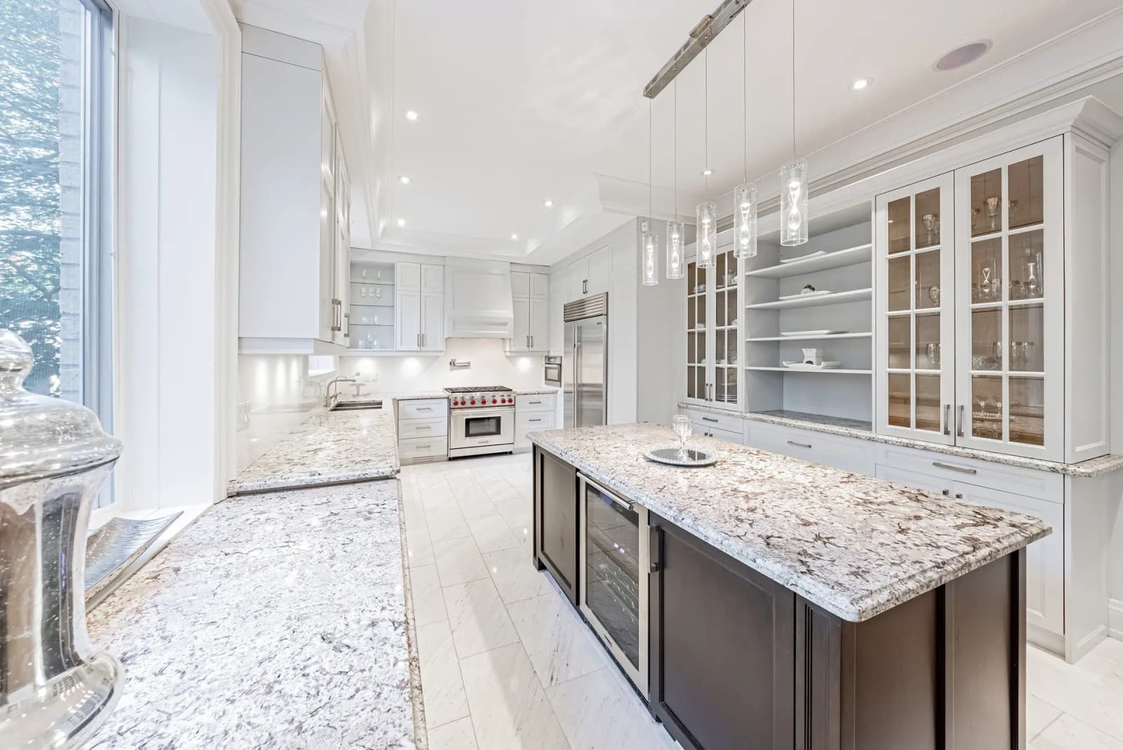 Transitional-looking Montréal kitchen, featuring a spacious central island with a veined granite countertop. The white cabinetry and brushed nickel handles add a touch of elegance to the space, while the neutral ceramic flooring creates a harmonious backdrop.