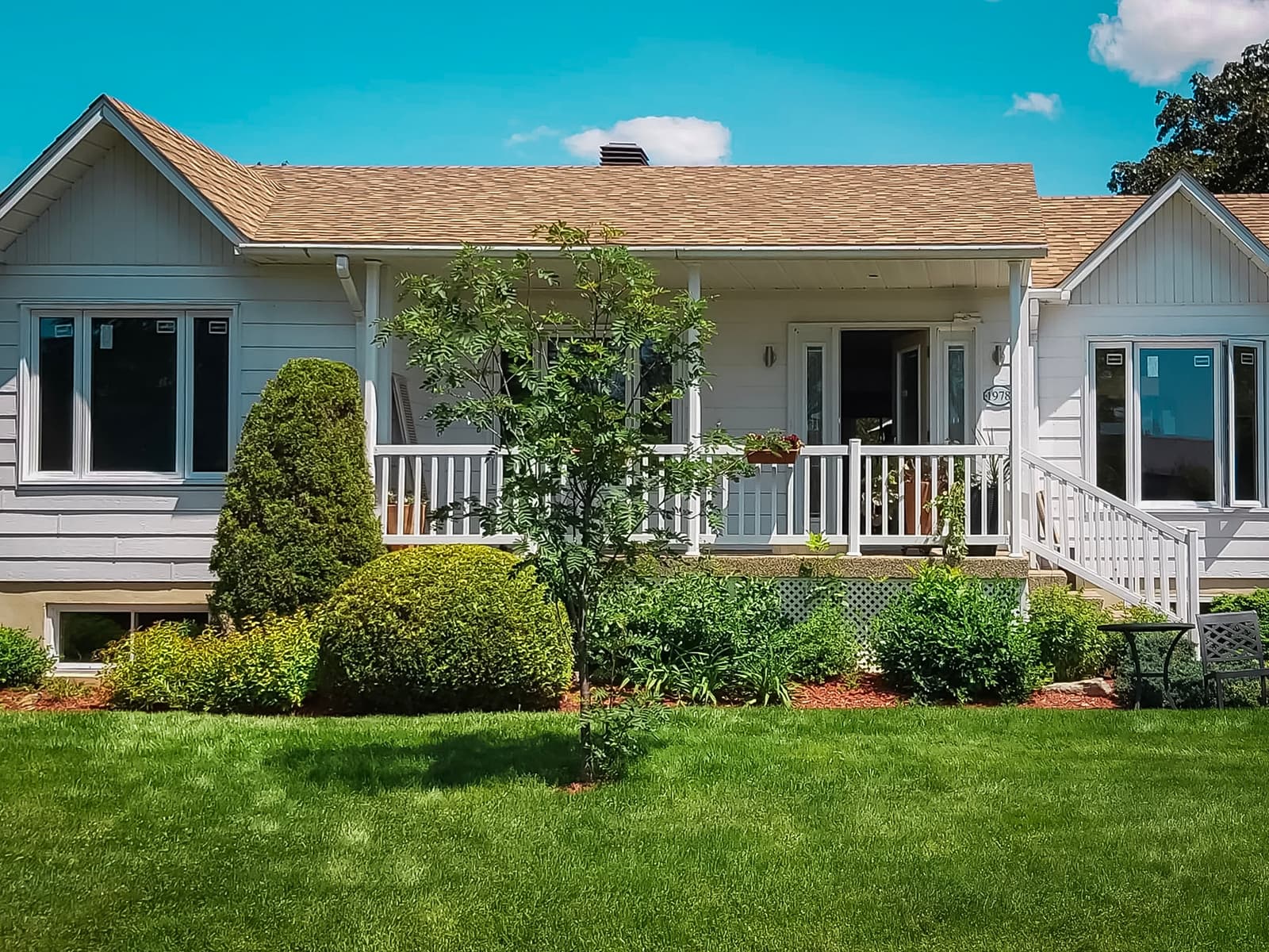 Single-story house with white wooden porch, landscaped garden, and green lawn under a clear blue sky.