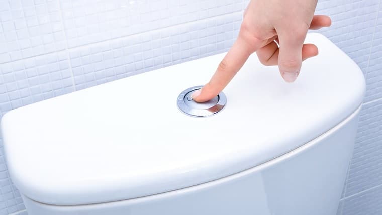 Hand pressing the flush button on a modern dual-flush toilet tank in a tiled bathroom.