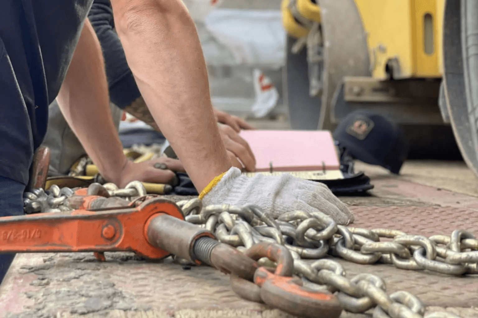 Worker handling lifting chains and tools on a construction site, with protective gloves and a binder of documents in view.