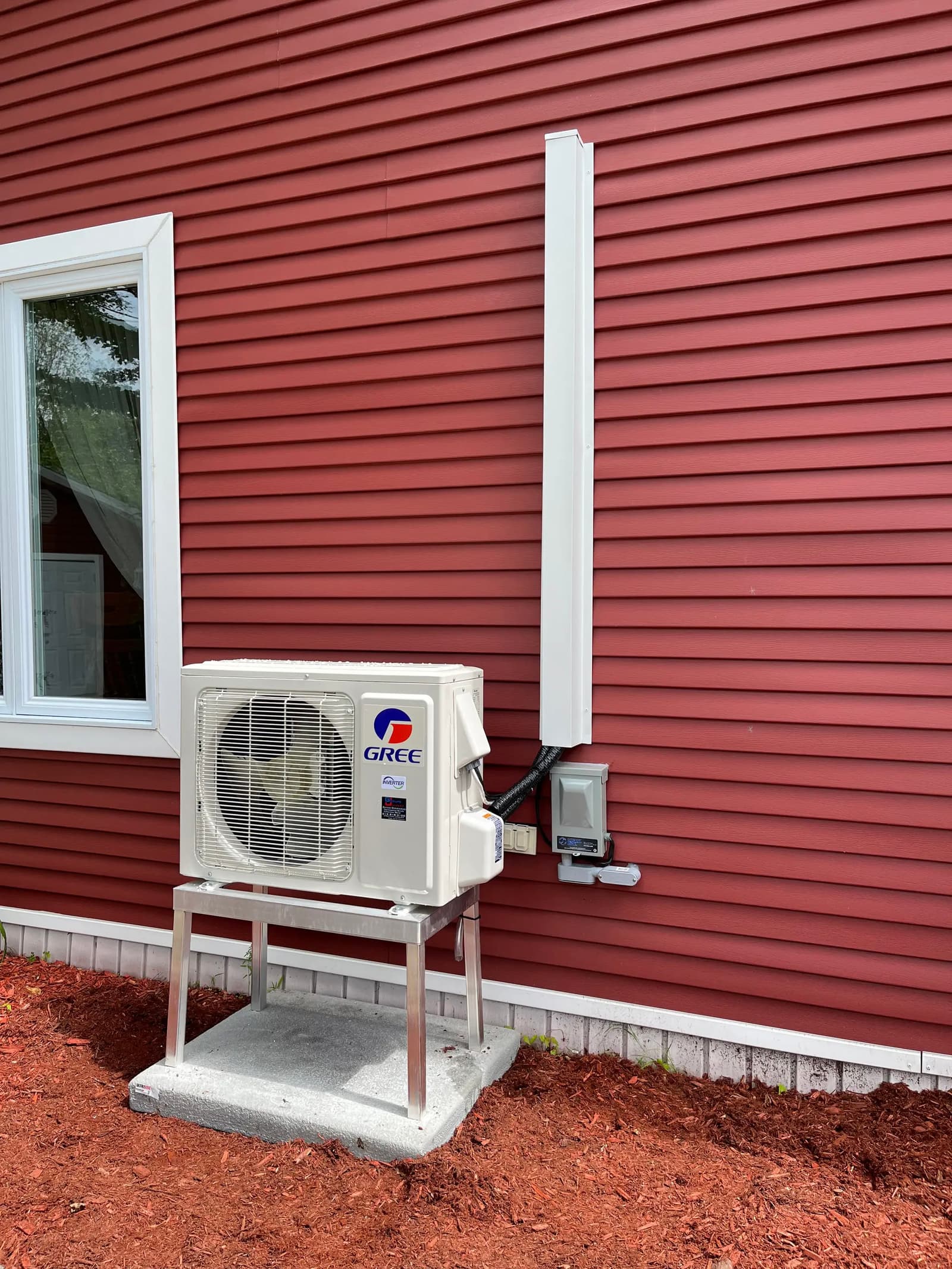 Outdoor Gree heat pump unit mounted on a red house wall with conduit casing and concrete base