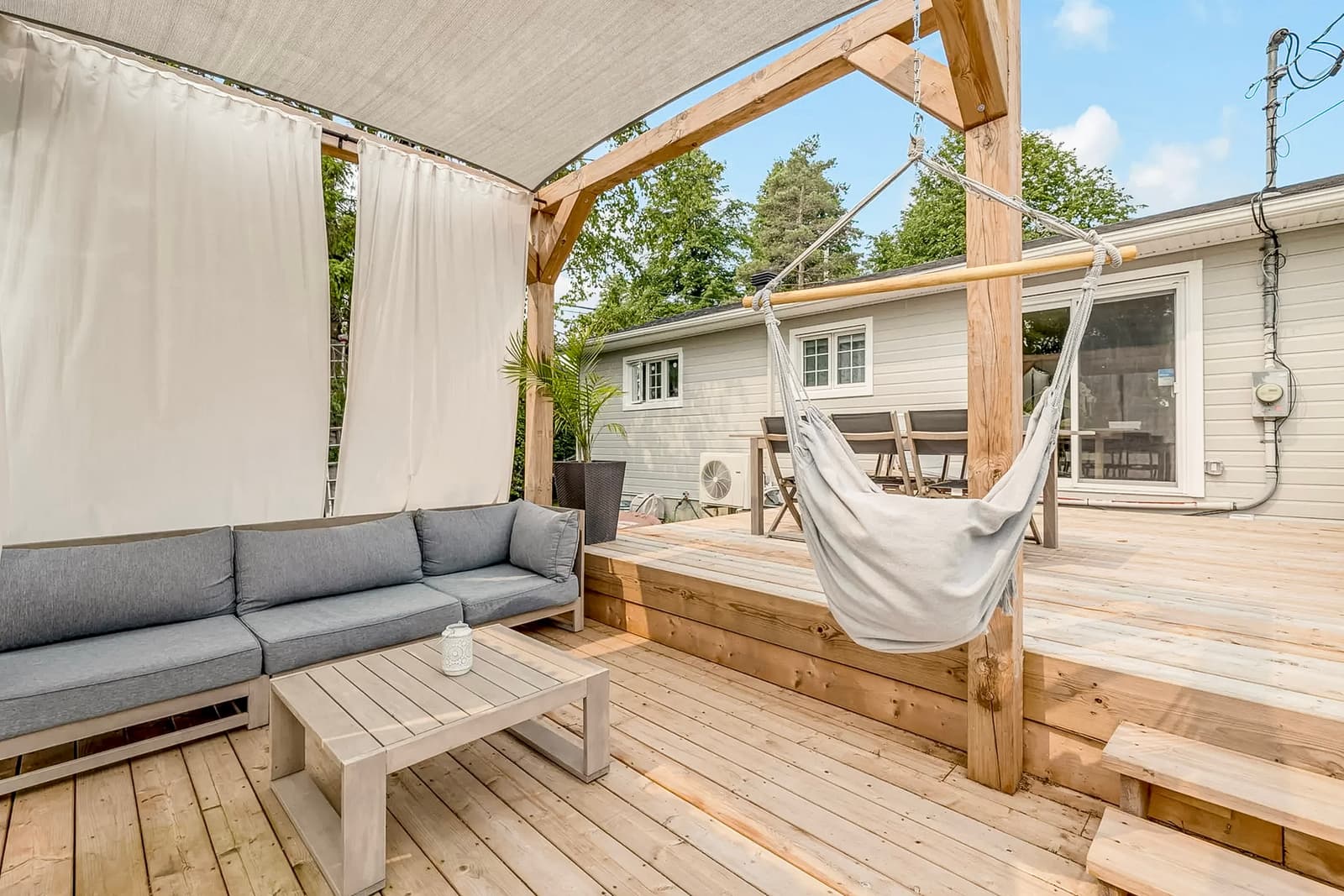 Wooden deck with lounge area, outdoor sofa, white curtains, and hanging chair under pergola