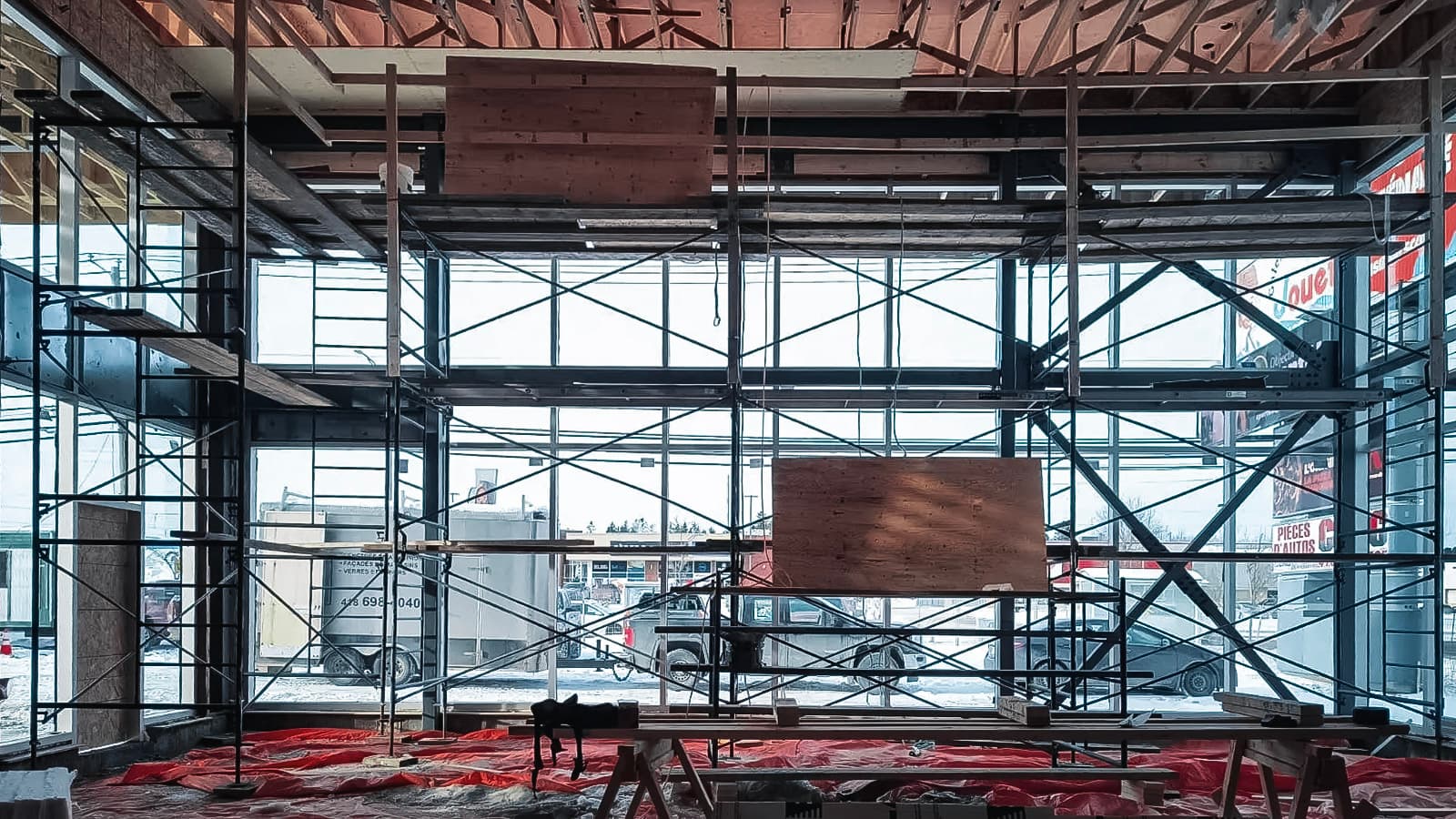 Interior construction site with metal scaffolding, large glass windows, wooden structures, and floor covered with a red tarp.