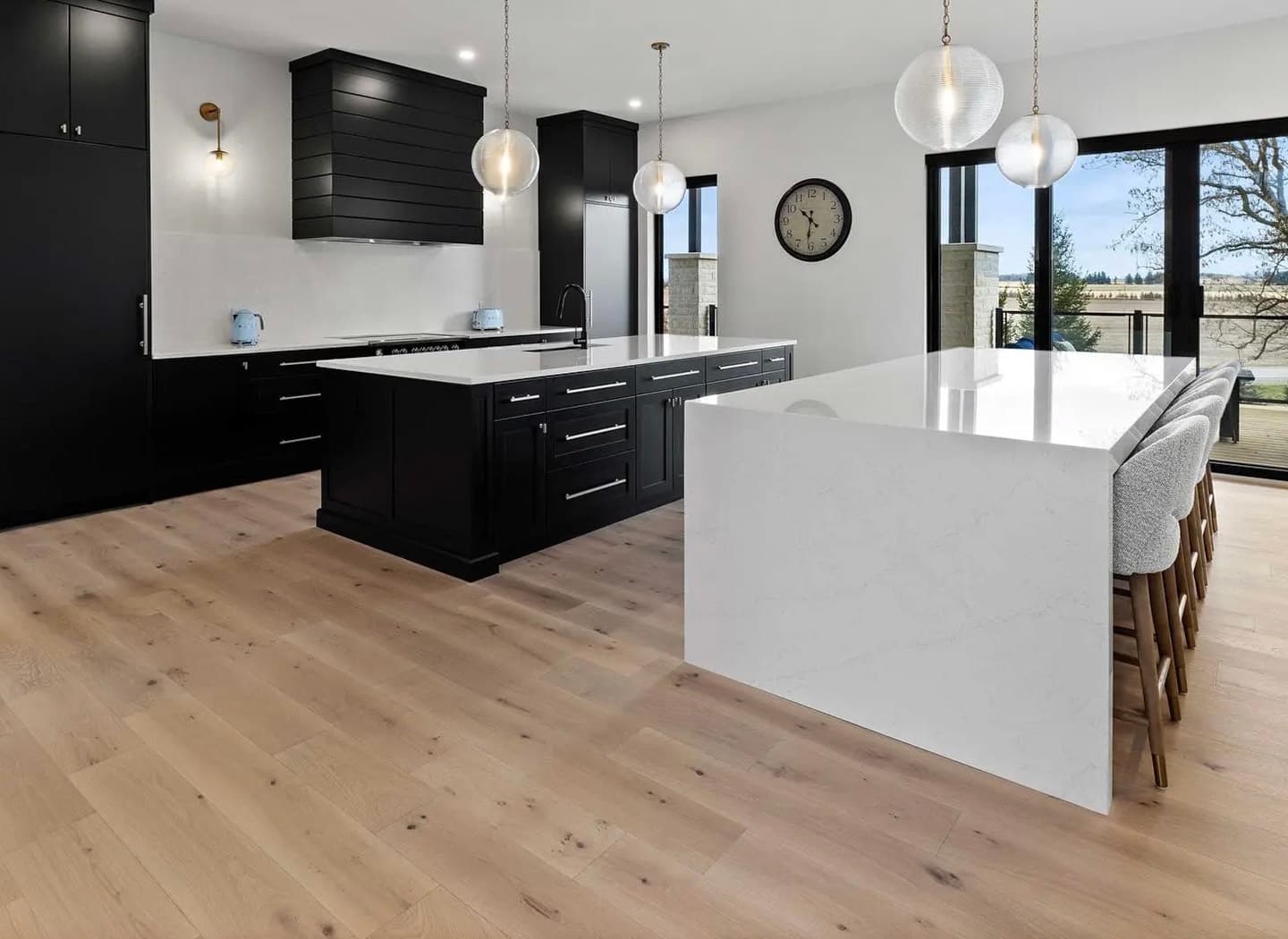 Modern kitchen with black cabinets, white marble island, and light wood flooring. Glass pendant lighting, large glass doors opening to the outside, and upholstered barstools.