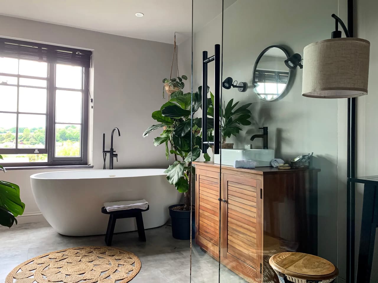 Bathroom with white freestanding bathtub, natural wood vanity, green plants, round mirror, and modern lighting.