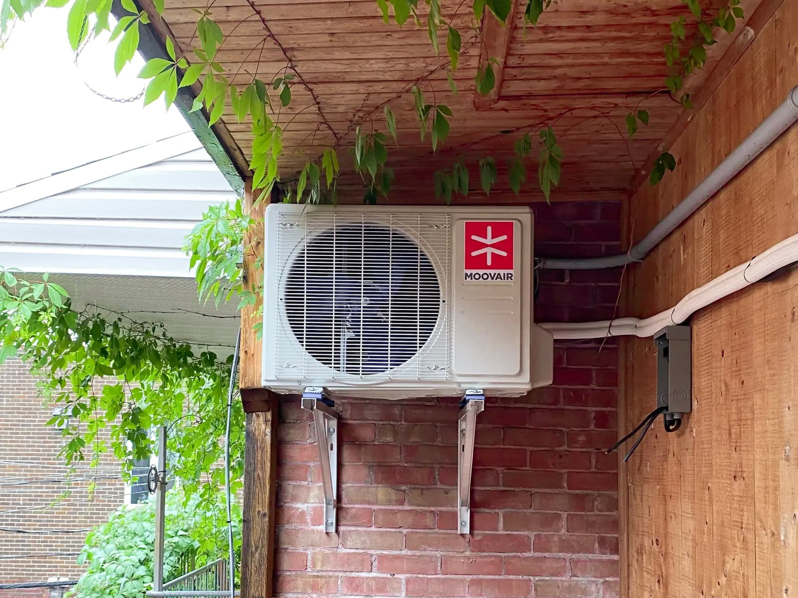 A Moovair heat pump outdoor unit mounted on a brick wall under a wooden roof, surrounded by vegetation, showcasing a harmonious and functional installation in a residential environment.