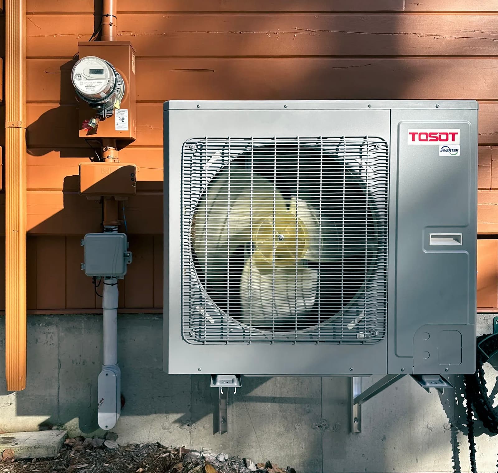 Tosot outdoor air conditioning unit mounted on an orange wooden wall with electrical meter and visible wiring.