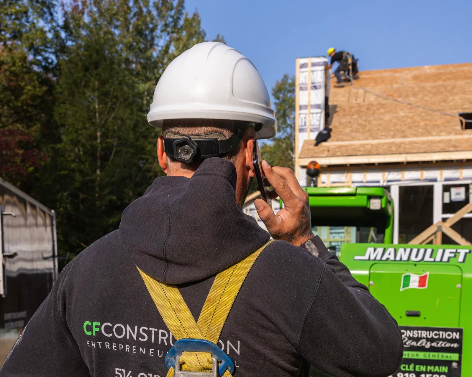 Construction worker wearing a safety harness and helmet, talking on the phone at a job site with roofing work in the background.