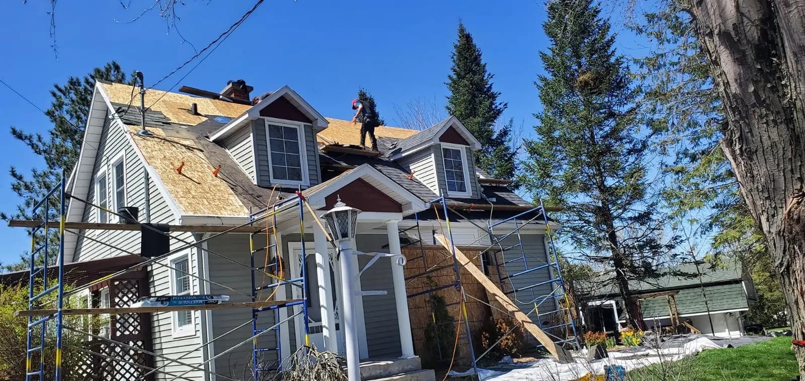 House under renovation with scaffolding surrounding it and workers on the roof installing plywood panels under a clear sky.