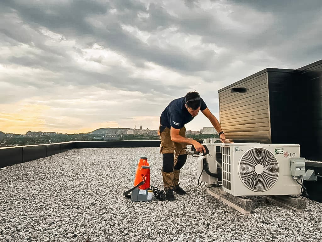 Technician installing an outdoor air conditioning unit on a flat roof with an urban view.