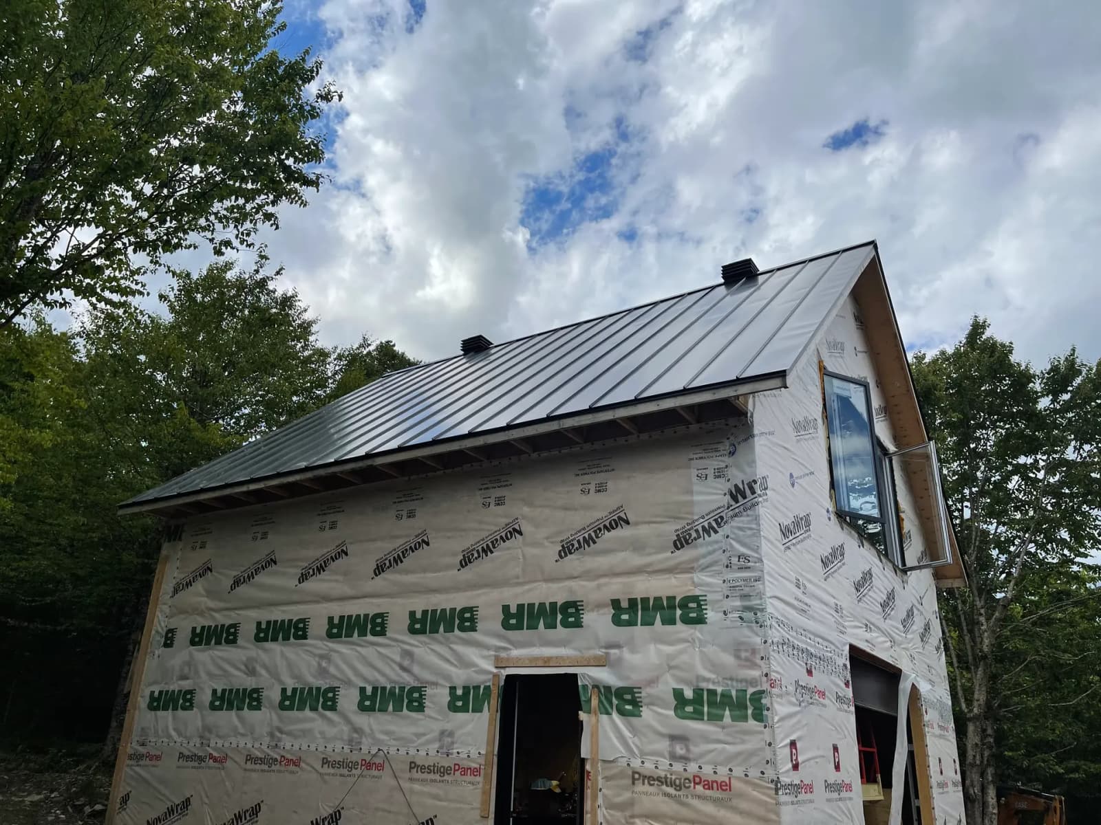 Residential building under construction, featuring a gray metal roof. The walls are still covered with protective materials and temporary insulation bearing the logos 'BMH' and 'PrestigePanel.' Set in a natural environment surrounded by trees, the structure has a gable roof design and an upper-floor window. The cloudy sky adds an outdoor construction site atmosphere.