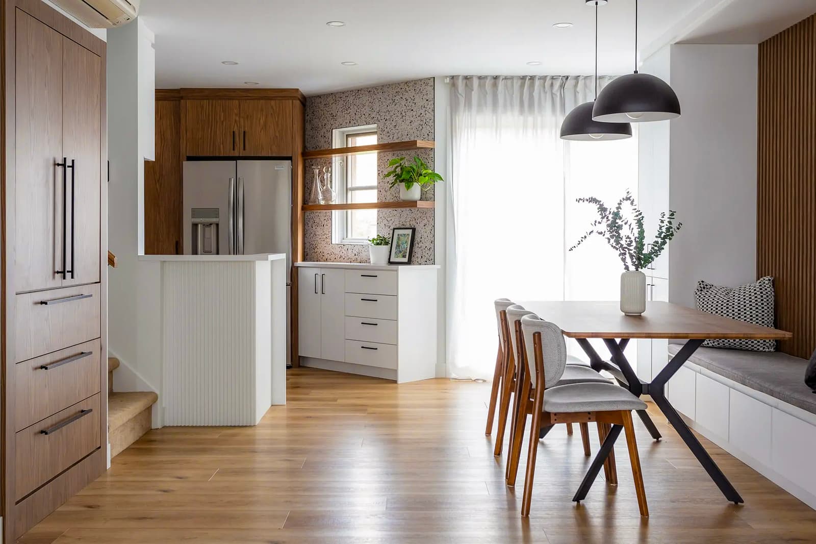 Modern kitchen and dining room with wooden furniture, wooden table with upholstered chairs, and built-in bench. Light wood flooring, black pendant lighting, and open shelves decorated with plants.Modern kitchen and dining room with wooden furniture, wooden table with upholstered chairs, and built-in bench. Black pendant lighting and open shelves decorated with plants.