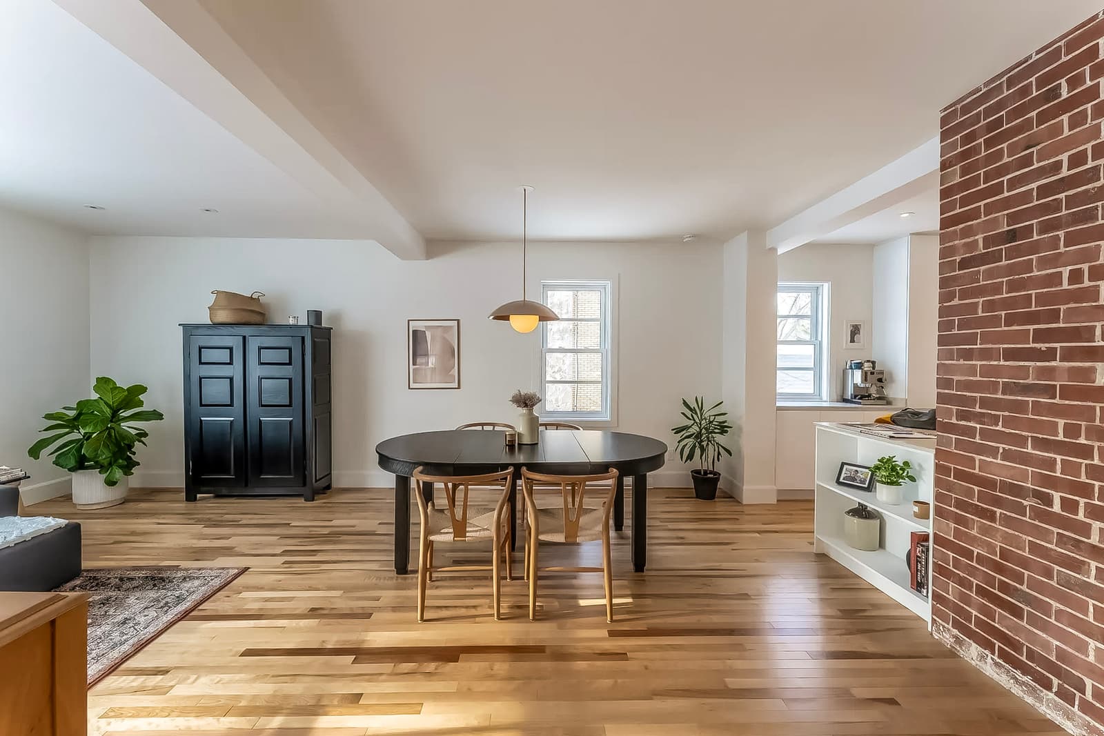 Modern dining room with a black table, wooden chairs, light hardwood flooring, and an exposed brick wall. Minimalist decor with indoor plants and pendant lighting.