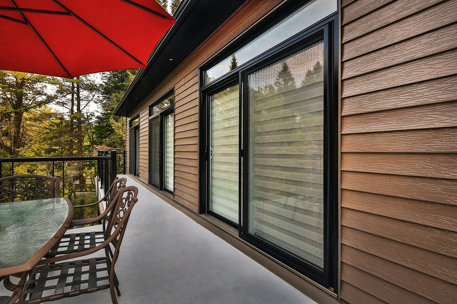 Modern outdoor terrace with wooden siding, large sliding windows, and metal patio furniture under a red umbrella.