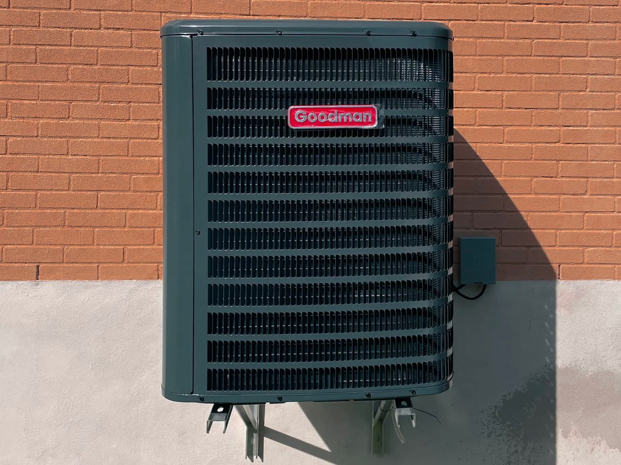 Black Goodman outdoor air conditioning unit mounted on an orange brick wall with concrete base.