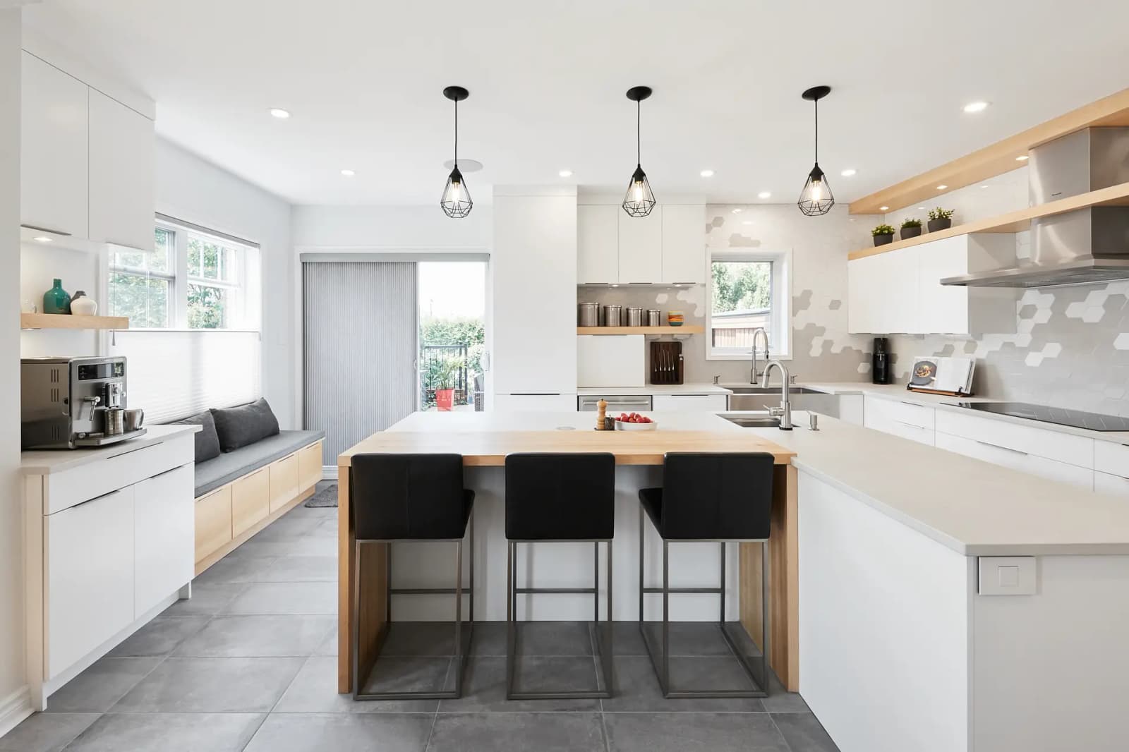 Scandinavian-style Montréal kitchen, featuring a spacious central island with black bar stools, minimalist white cabinets and a geometric mosaic backsplash. The grey ceramic floor tiles add a touch of warmth to the space.
