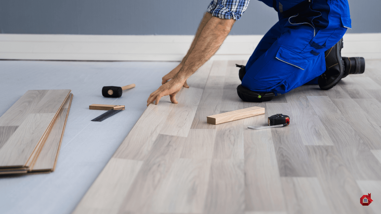Installation of light-colored floating flooring by a kneeling renovator, with visible tools (mallet, ruler, spacers, measuring tape)
