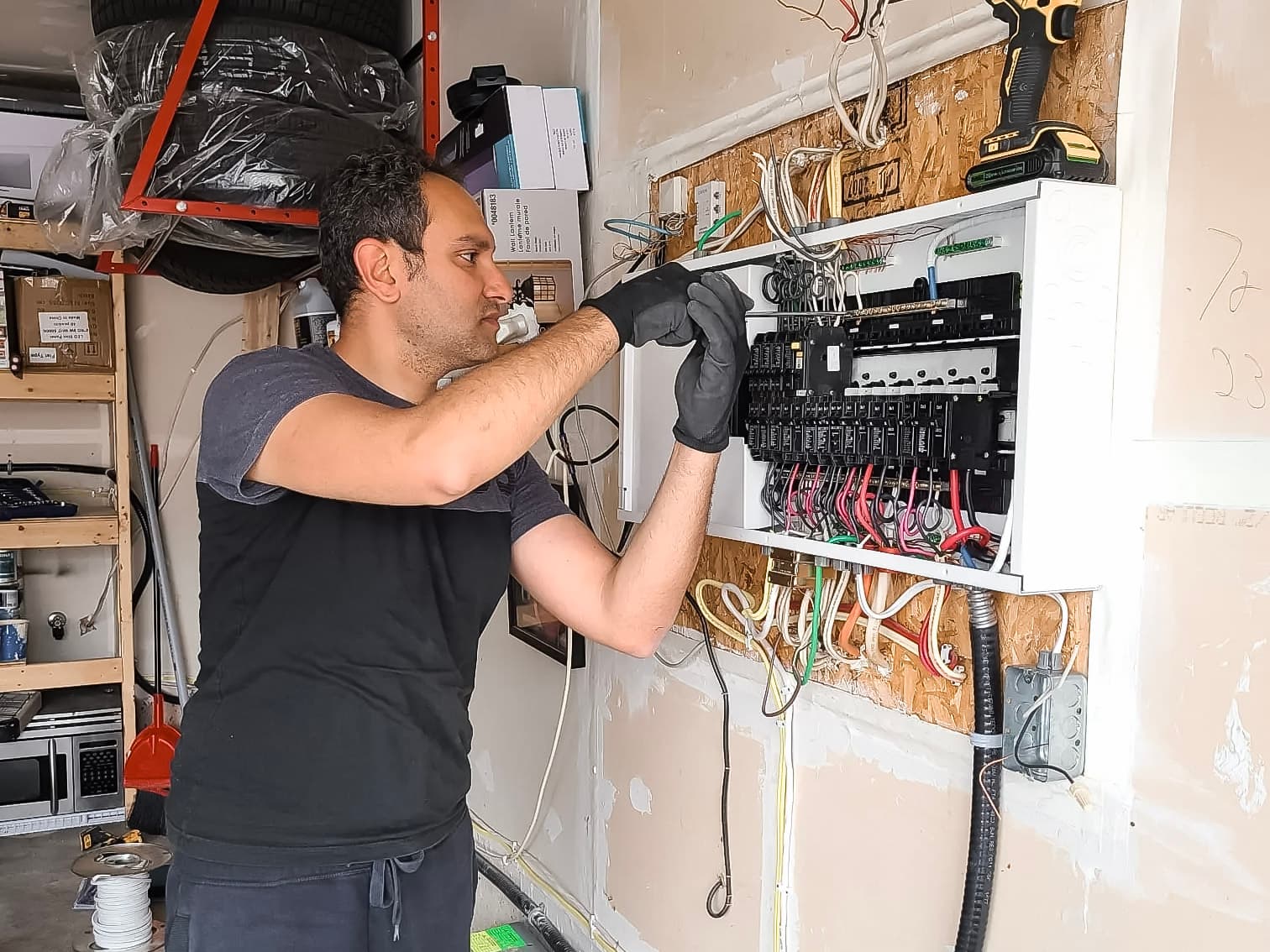 An electrician working on a circuit breaker panel in a garage, with exposed wiring and tools.