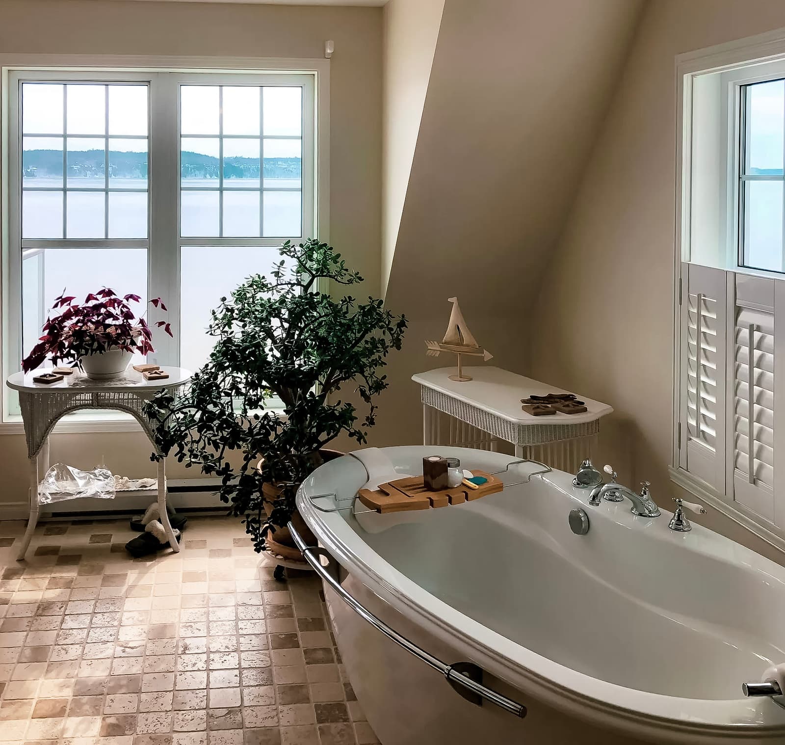 Bright bathroom with freestanding bathtub, natural stone tile flooring, and large windows overlooking an outdoor landscape. White wicker furniture and soothing plant decor.