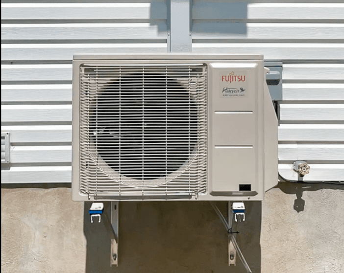 Fujitsu Halcyon outdoor heat pump unit installed on a white wall of a residential house, mounted on a metal bracket for a modern HVAC system, providing heating and cooling with optimal energy efficiency, ensuring thermal comfort.