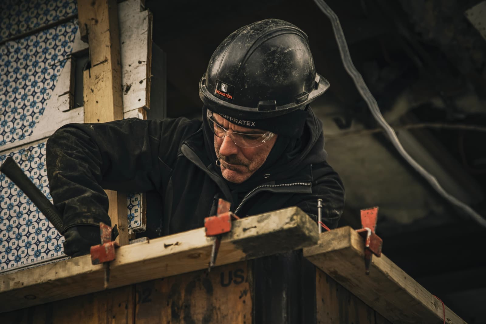 Worker wearing a black helmet and safety glasses, performing carpentry and construction tasks on a job site.