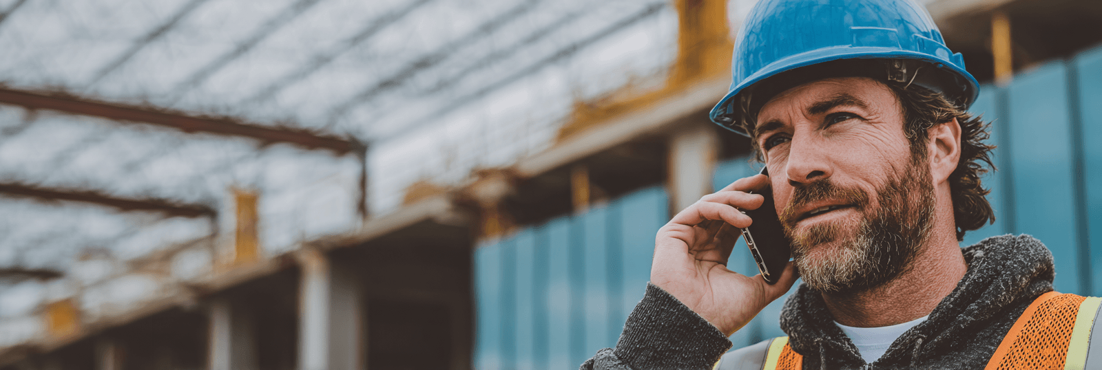 Construction contractor wearing a blue hard hat and high-visibility vest while talking on a phone at a job site