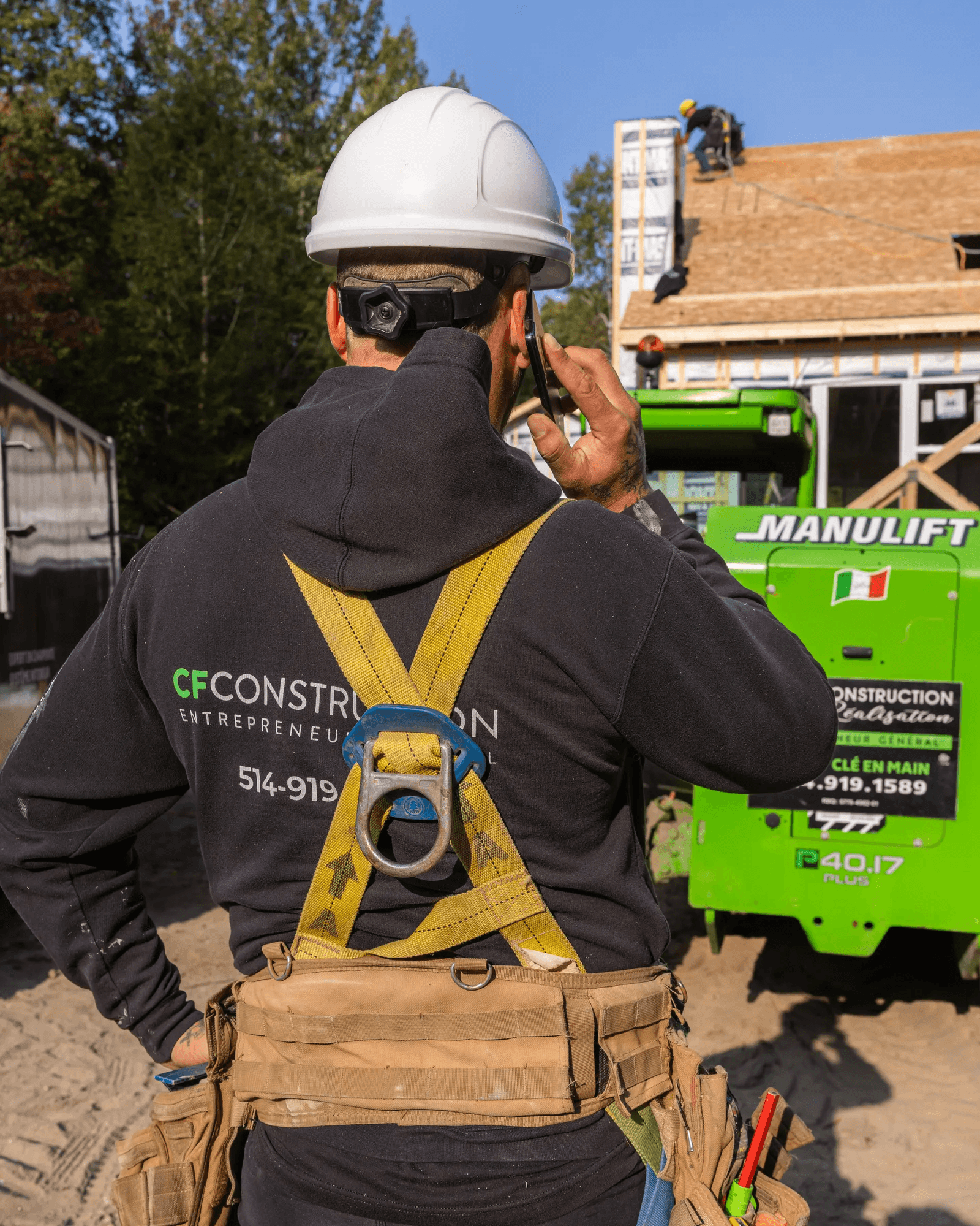 Construction worker wearing a safety harness and helmet, talking on the phone at a job site with roofing work in the background.