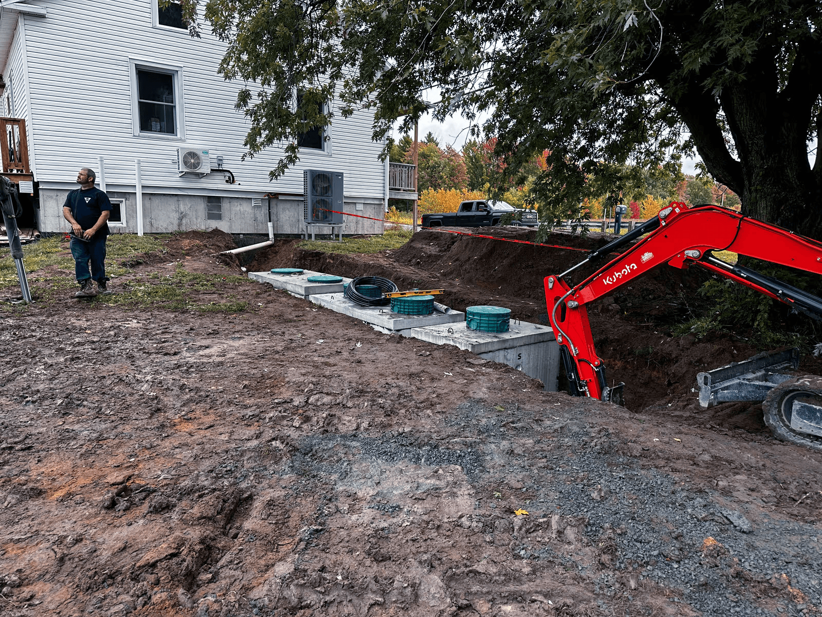 Residential septic system installation beside a house with excavation, concrete septic tanks, access risers, piping, and a compact excavator on site.