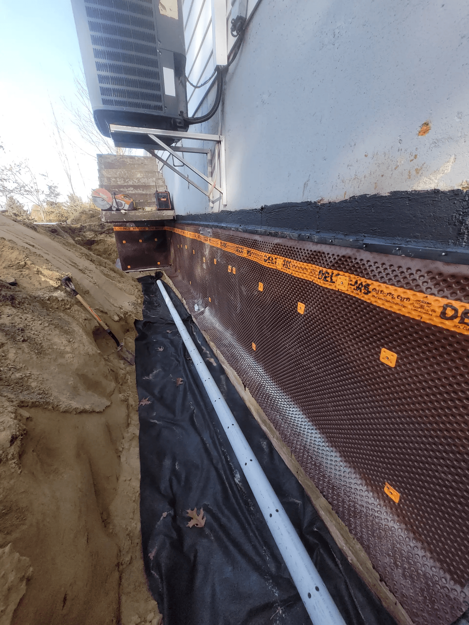 Exterior French drain installation along a foundation with drainage membrane, perforated pipe and excavation around the house