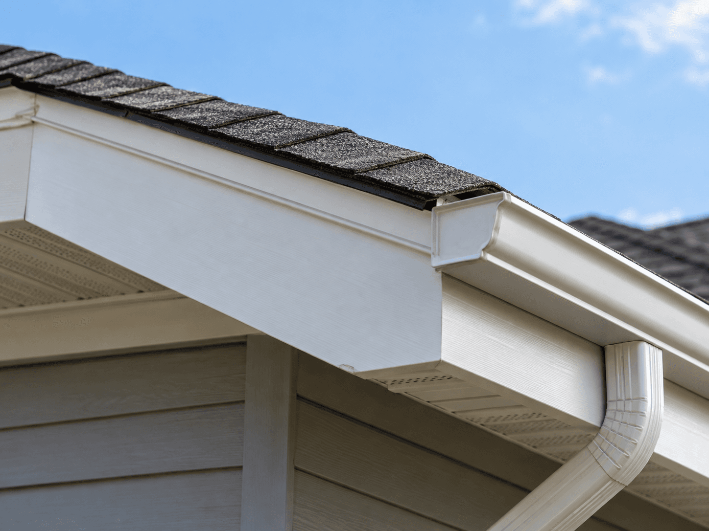 Close-up of a residential roof with dark grey asphalt shingles, white gutter, downspout, white fascia boards and perforated ventilation soffits beneath the eaves of a house with grey exterior siding