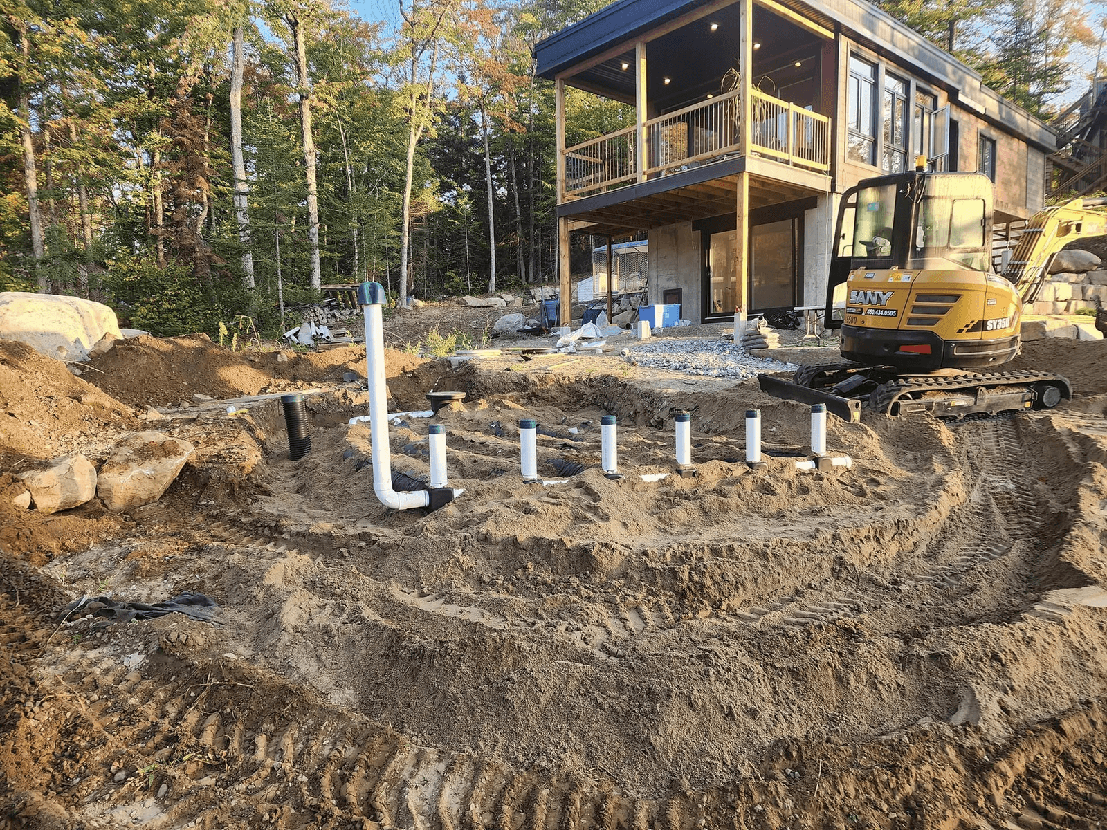 Residential septic system installation beside a house with excavation, distribution piping, multiple access risers and a compact excavator preparing the site.