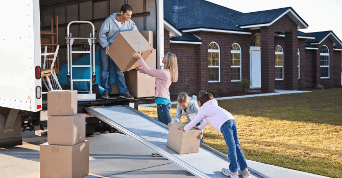 A family unloads cardboard boxes from a moving truck outside a brick house.