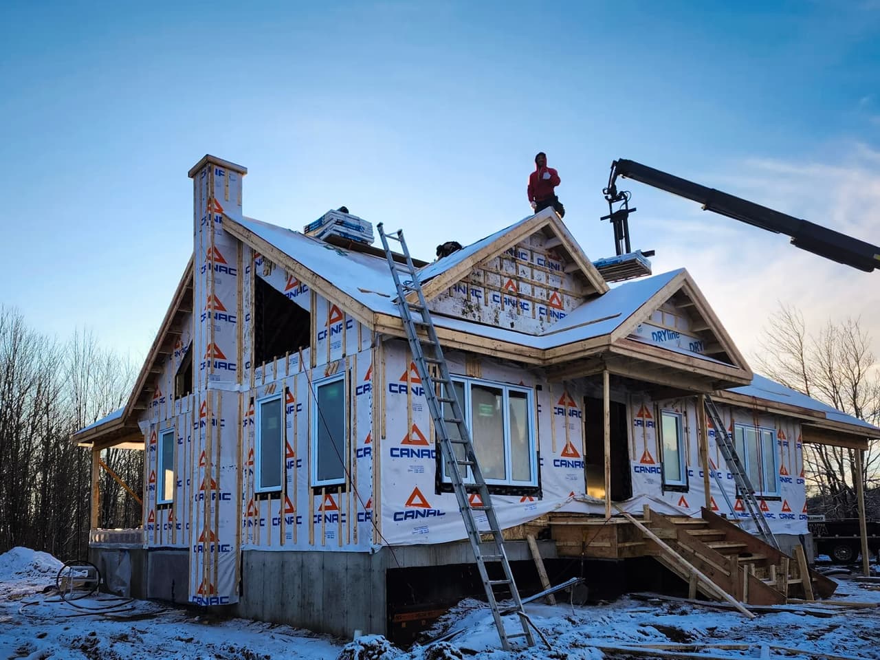 House under construction in winter with CANAC weather barrier wrap, scaffolding, ladders, and workers on the roof under a blue sky.