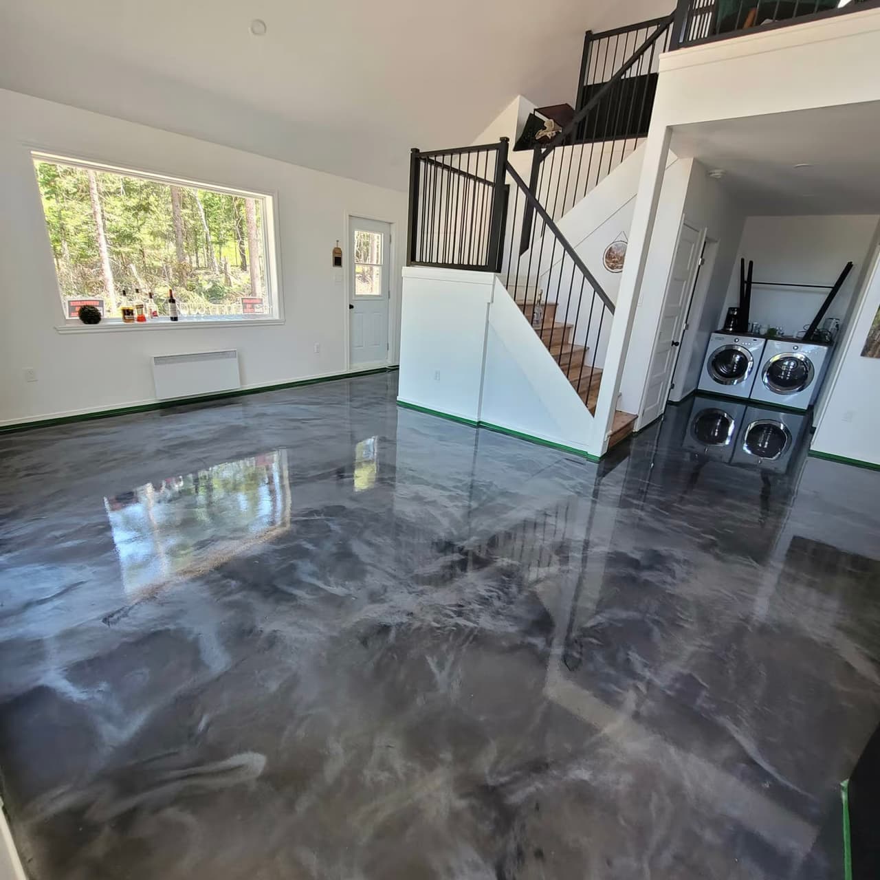 Large interior room with glossy gray epoxy flooring, staircase with black railings, mezzanine, panoramic window and laundry area.