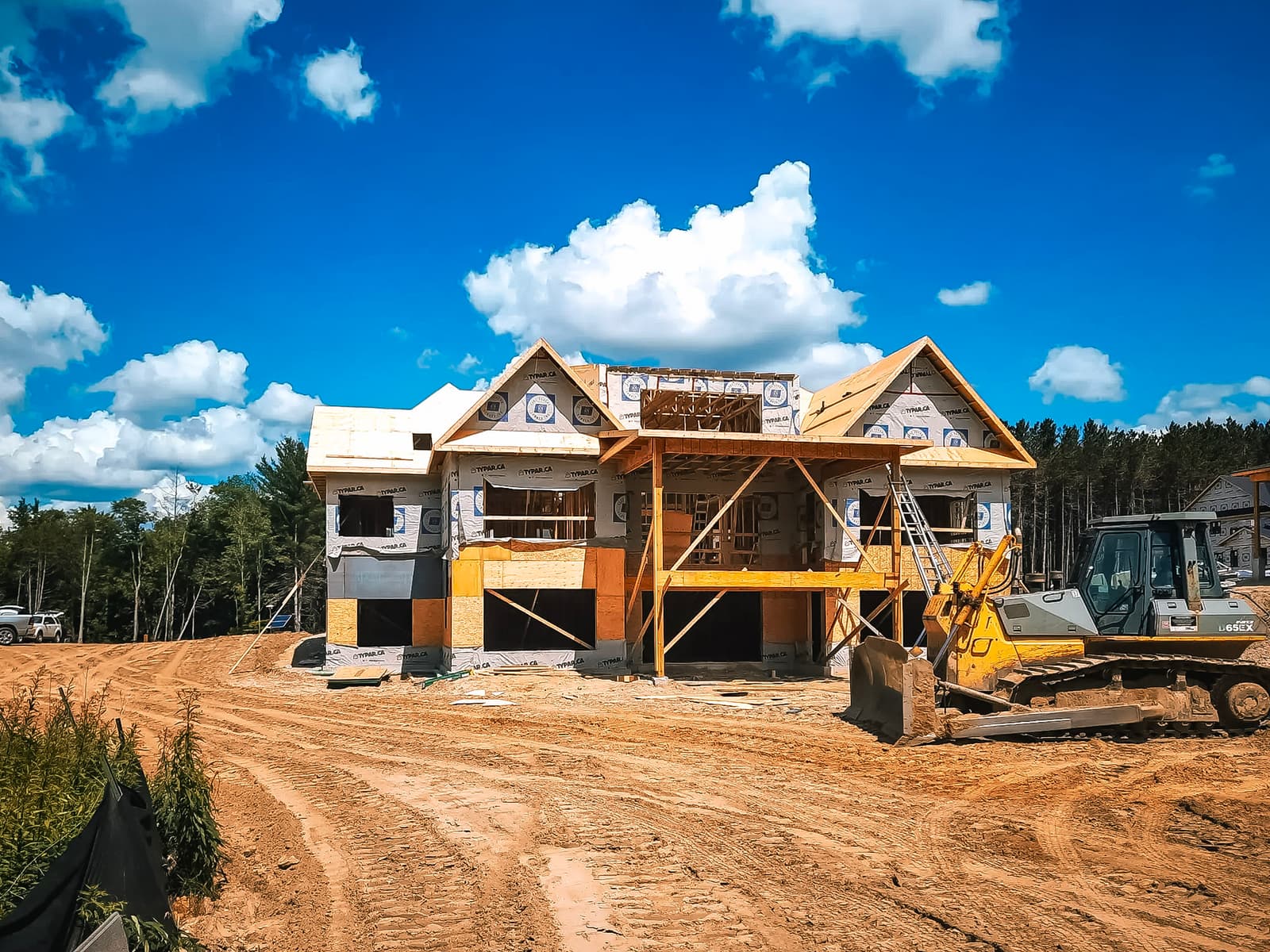 Two-story house under construction on a sandy lot with Tyvek weather wrap, wooden scaffolding, and heavy machinery on site.