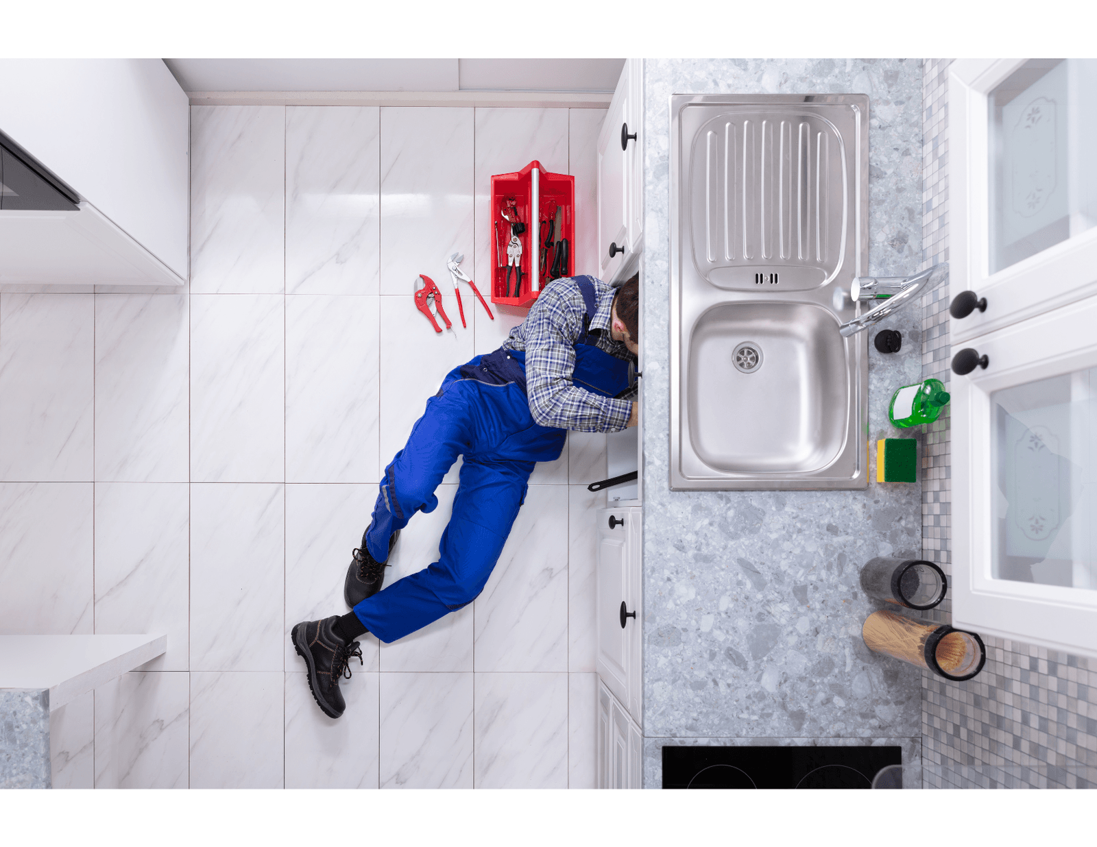 Plumber in blue overalls repairing the plumbing under a modern kitchen sink with a toolbox nearby.