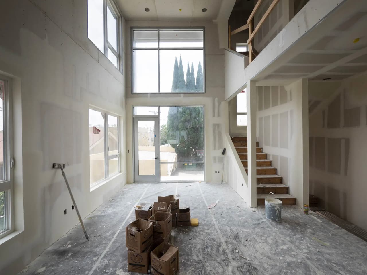 House interior under construction with cathedral ceiling, mezzanine, staircase, drywall walls, and large windows allowing abundant natural light.
