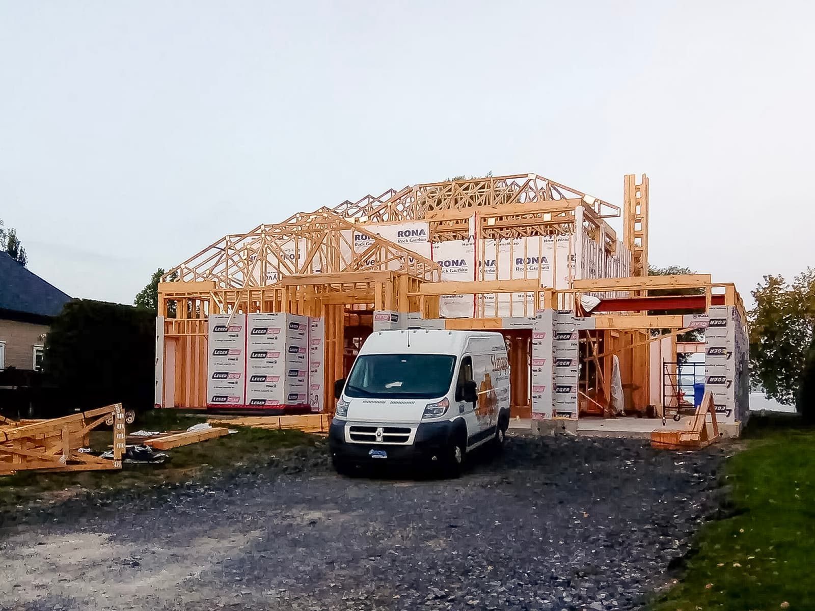 House under construction with wooden framing, RONA weather wrap, and a work van parked on the gravel driveway.