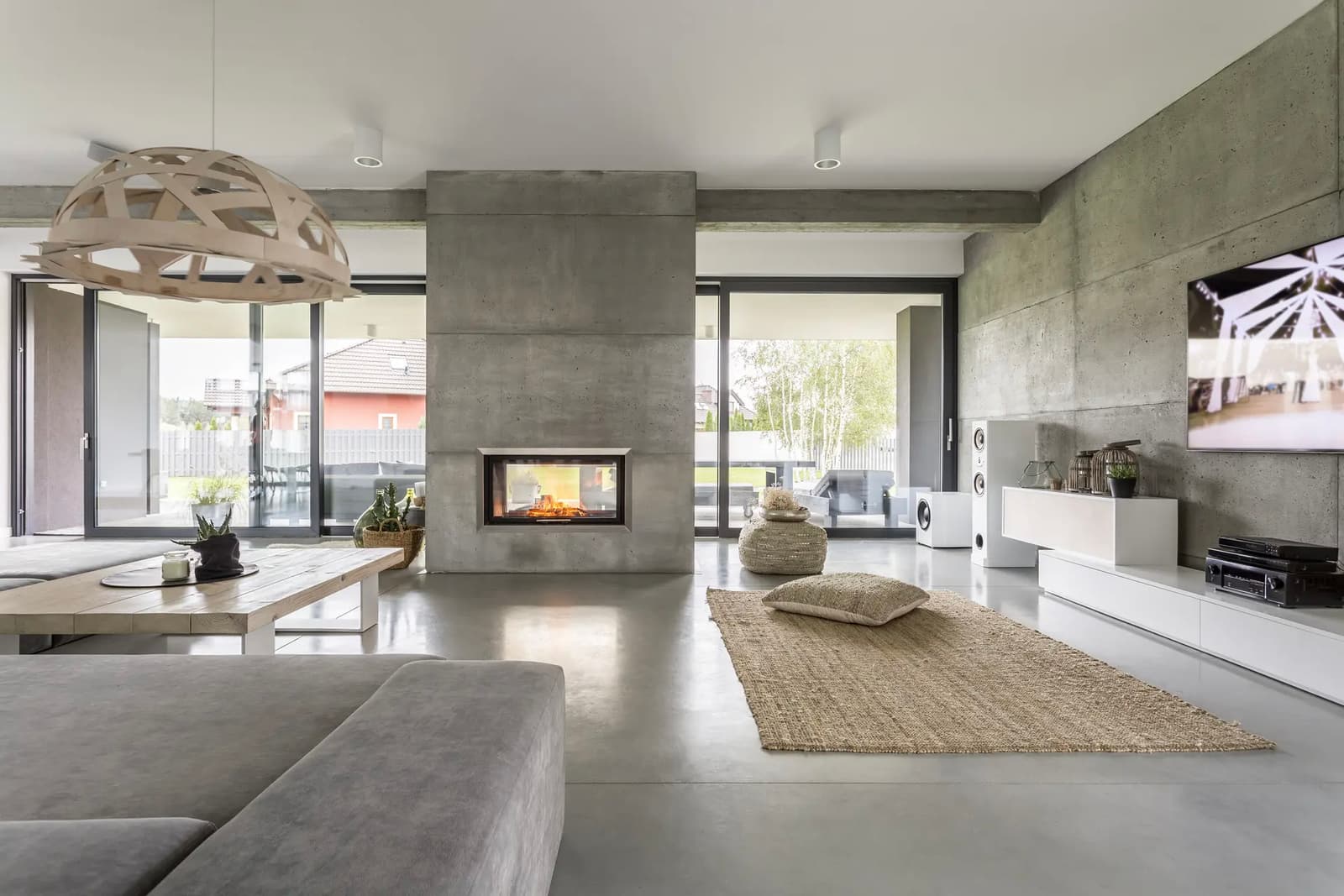 Modern living room with an industrial style featuring raw concrete walls, polished concrete flooring, a built-in fireplace, and large glass doors opening to an outdoor terrace.