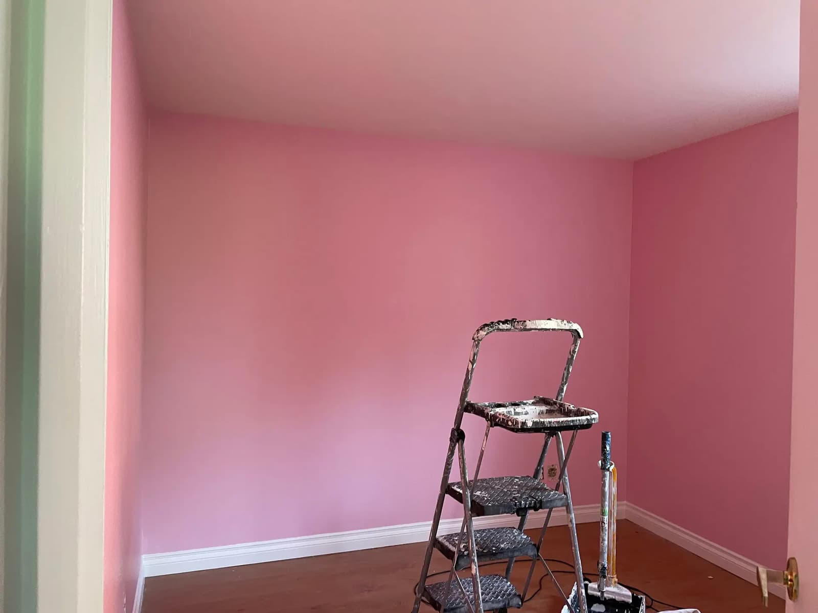 Room under renovation with freshly painted pink walls, white baseboards, wooden floor, and painting equipment.