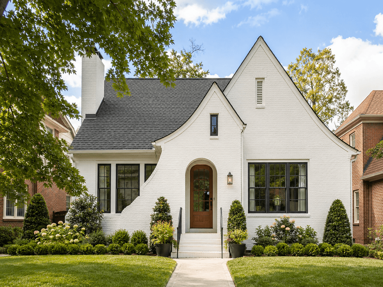Elegant white house with a painted brick façade, dark roof, black-framed windows, an arched entrance, and manicured landscaping.