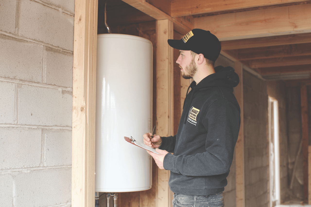 A technician wearing a black cap and hoodie inspects a white water heater in a house under construction, taking notes on a clipboard.