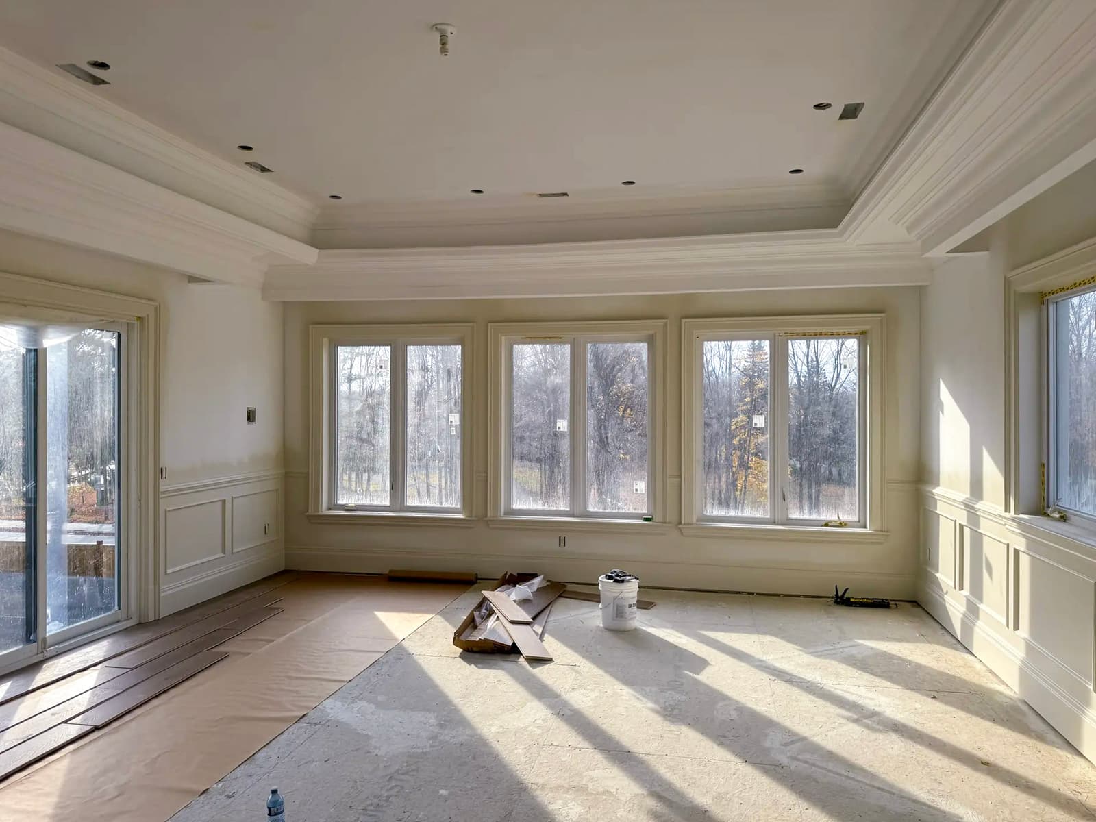 Bright room under renovation with decorative moldings, large windows, coffered ceiling, and flooring under installation.