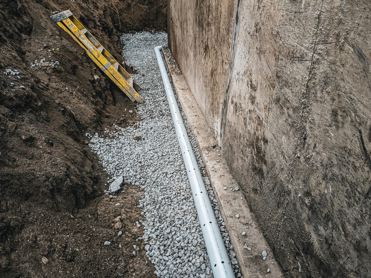 Installation of a perforated PVC French drain with gravel along the foundation of a house.