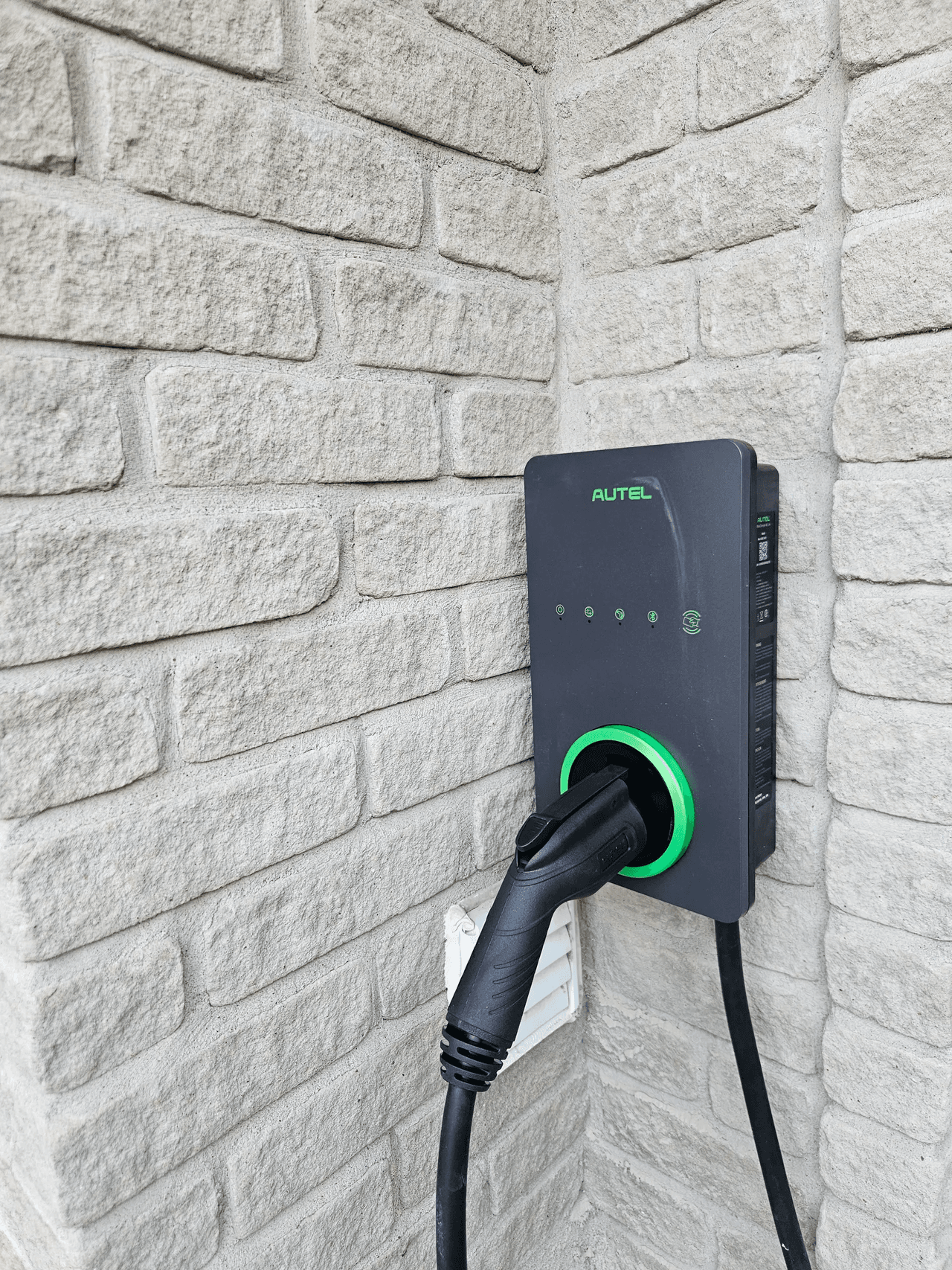 Electric vehicle charging station installed on a light brick exterior wall, with the charging cable plugged in.