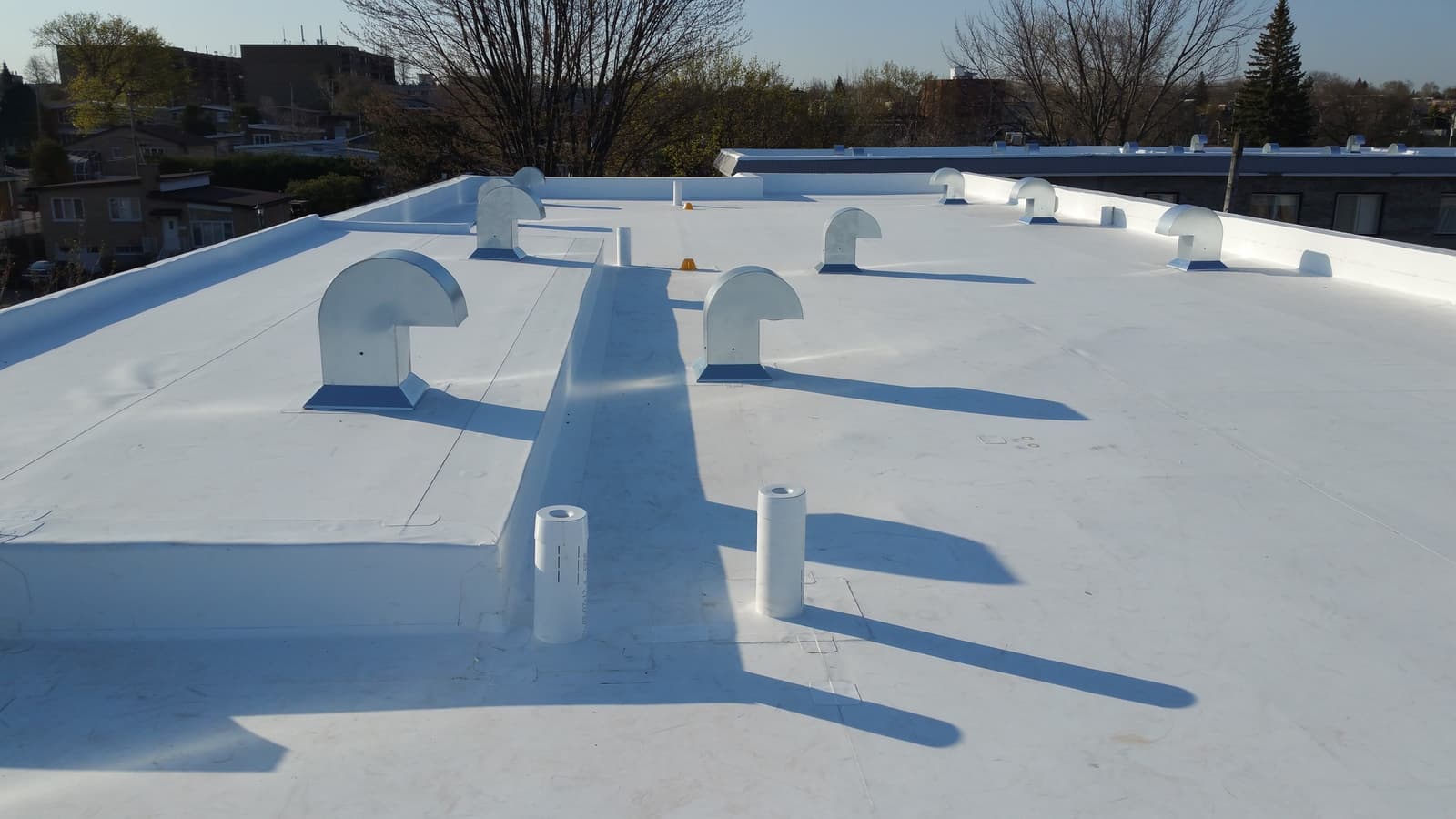 Flat roof covered with a white membrane featuring multiple metal vents and drains, ensuring waterproofing and ventilation for a residential building.